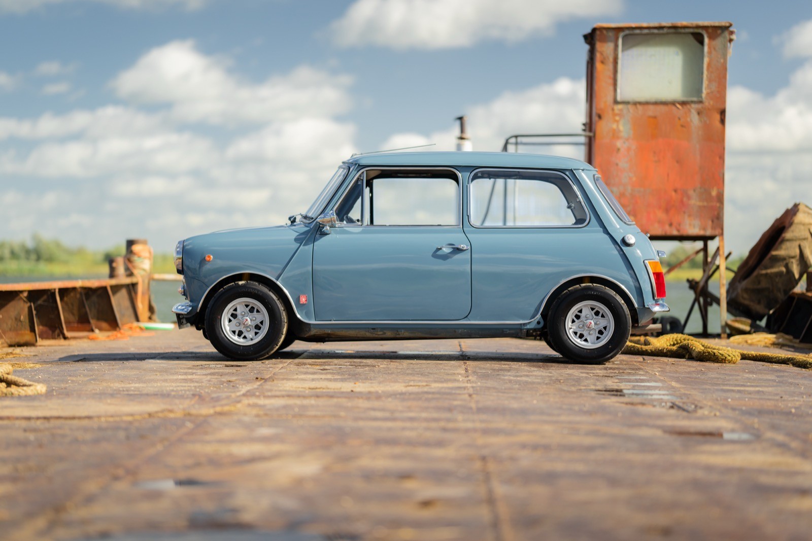 A light blue vintage car parked beside an old building under a cloudy sky, with rusted objects nearby.