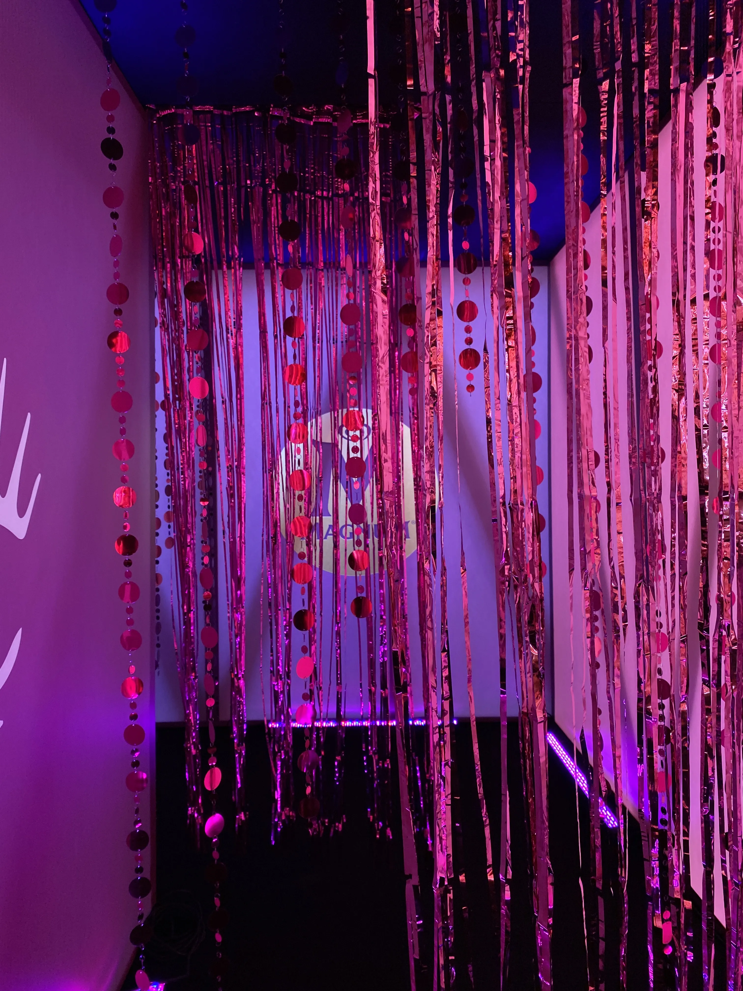 Shimmering tinsel curtain installation in pink and purple light with glowing logo in the back.