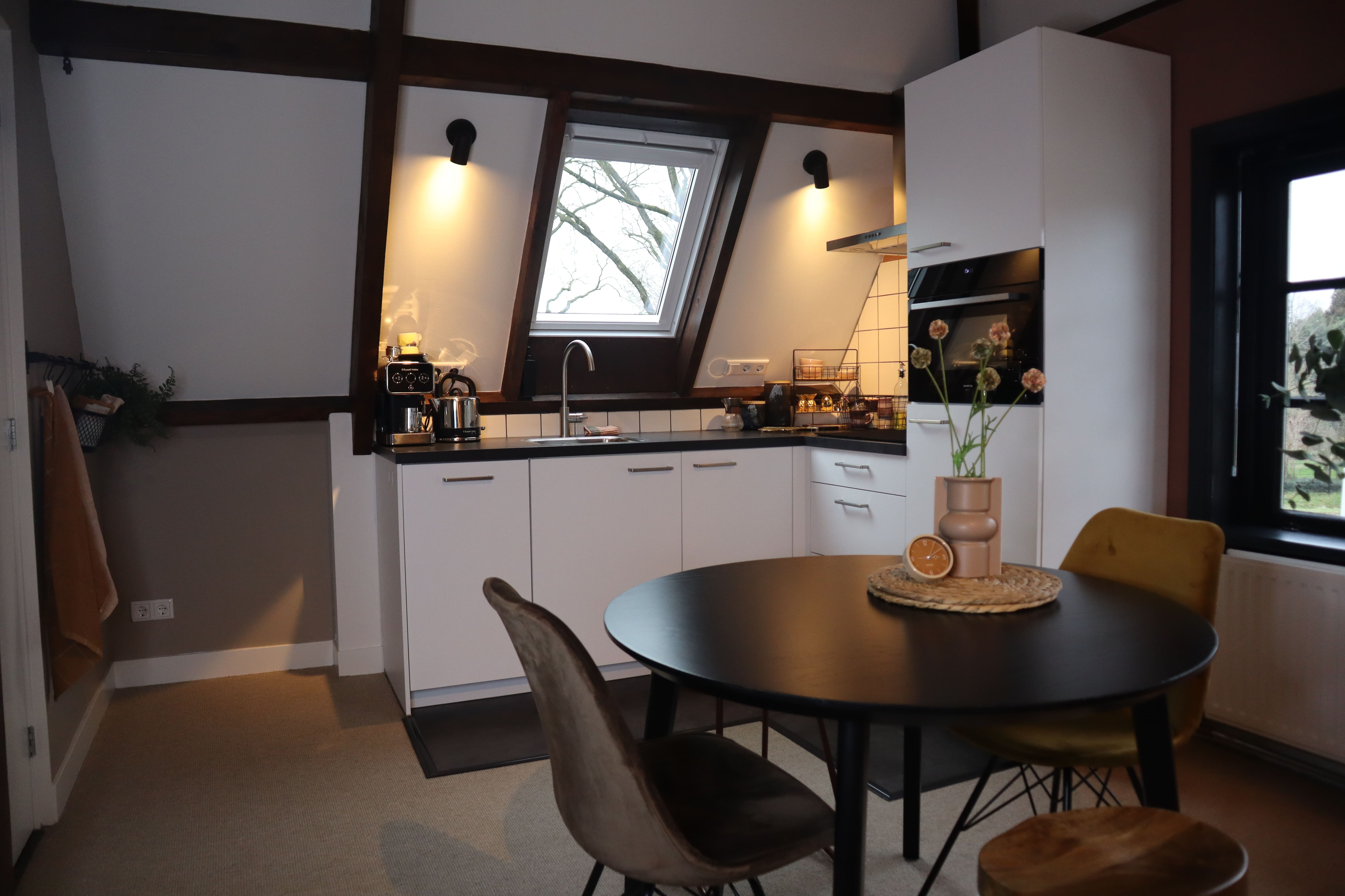 Kleine keuken met witte kasten, een fornuis en een eettafel met stoelen. Het raam biedt uitzicht op de buitenomgeving en de kamer heeft houten balken.
