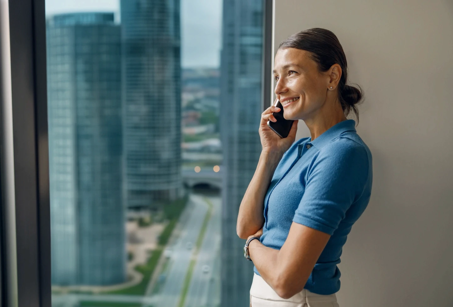 A smiling woman in a blue polo shirt is standing by a window, talking on a smartphone with modern city skyscrapers visible in the background, reflecting a professional and urban environment.
