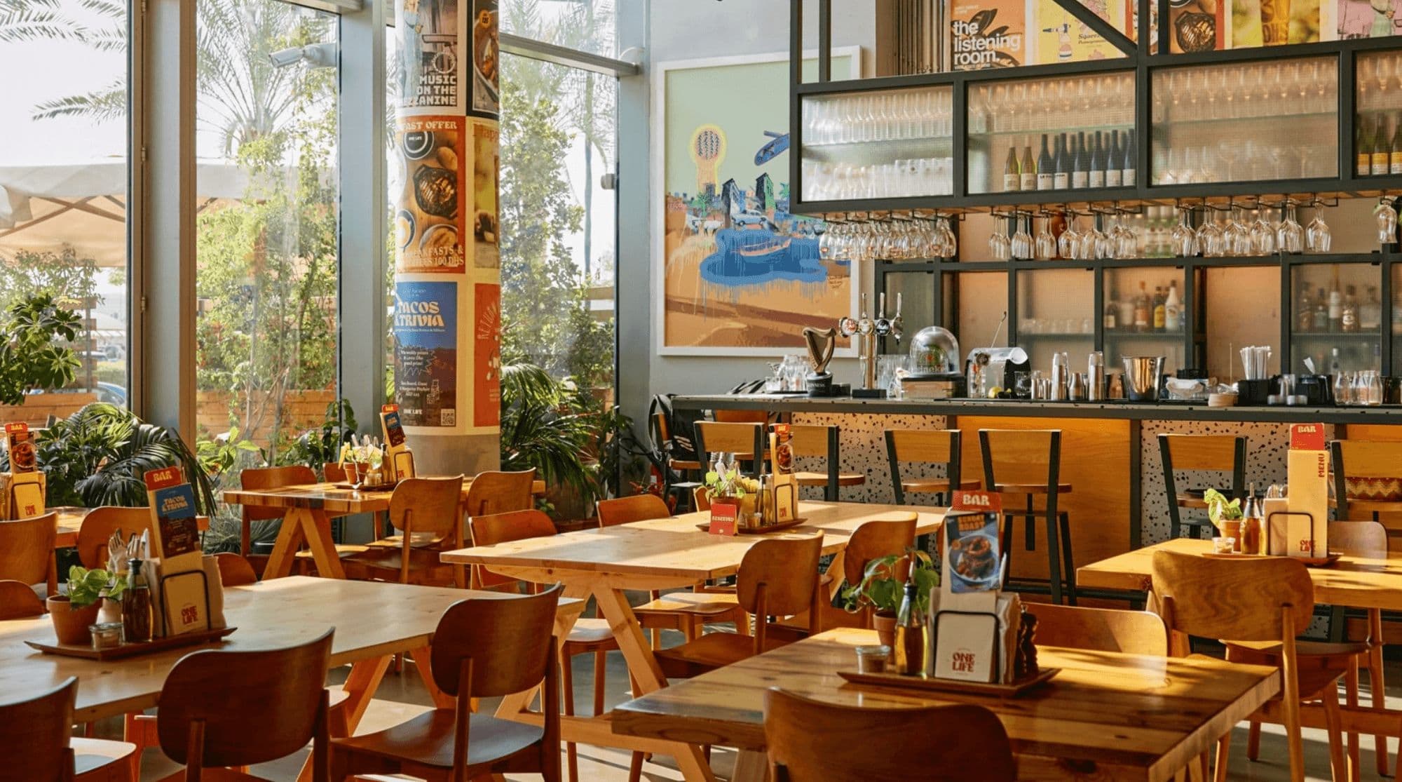 OneLife Kitchen in Dubai with an open bar, wooden seating and tables, floor-to-ceiling windows, and greenery.