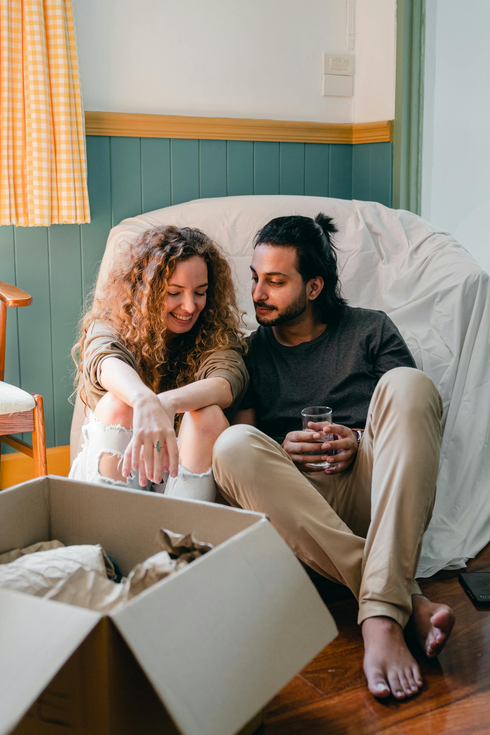 A happy couple sits barefoot on a wooden floor in a partially unpacked room. The woman, with long curly hair, is laughing, and the man is smiling at her while holding a glass of water. They are sitting in front of a chair draped in a white sheet, with an open box in the foreground.