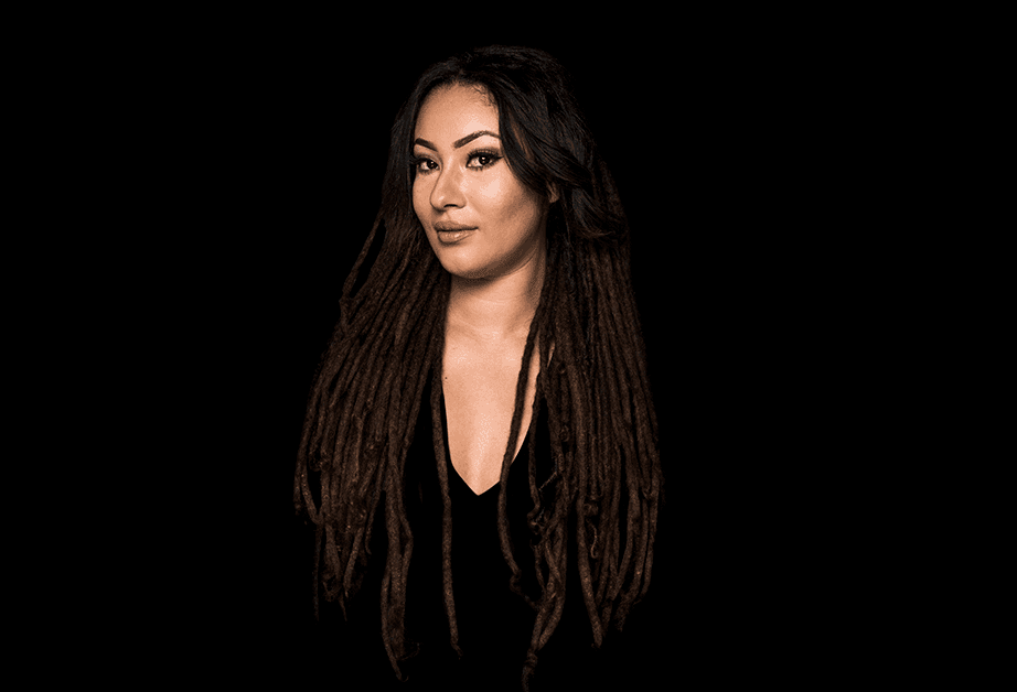 A woman with long, dark dreadlocks stands against a solid black background. She wears a black top with a deep V-neck. Her gaze is directed slightly to her left, and her expression is neutral. The lighting highlights her face and hair, creating a contrast against the dark backdrop. No other objects or text are visible in the image.