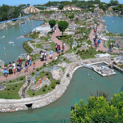 Aerial view of a miniature park with model buildings, pathways, water features, and visitors walking and observing the miniatures.