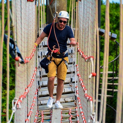 Un hombre con casco y arnés camina por un puente de cuerda con redes, rodeado de frondosos árboles verdes.