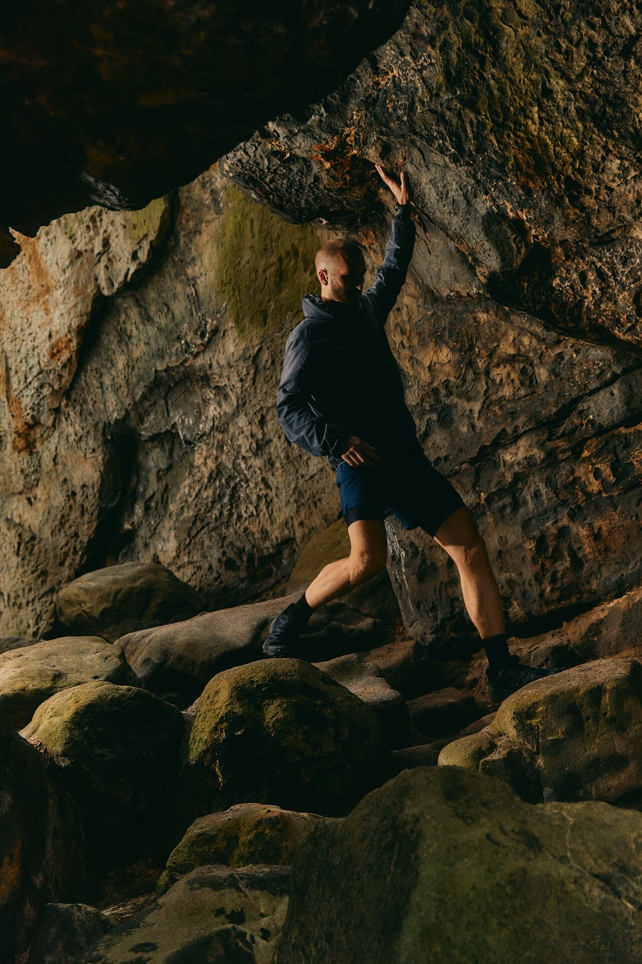 A person stands confidently on moss-covered rocks in a dimly lit cave, touching the rocky ceiling, conveying a sense of adventure and exploration.