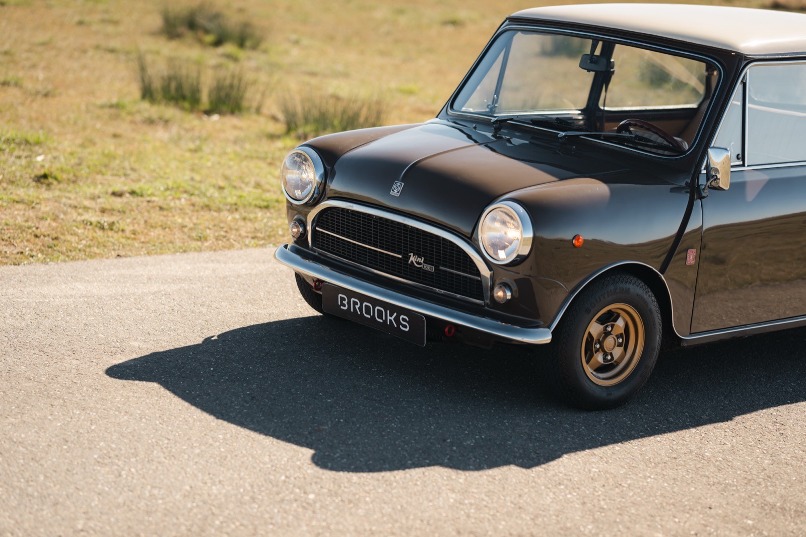 A classic small car parked on a road with a grassy background, showcasing its vintage design and shadows.