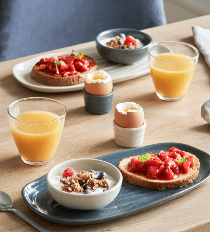 Table en bois dressée pour deux petits déjeuners avec plateaux en grès bleu, bols de granola, tartines aux fraises, œufs à la coque dans des coquetiers en céramique et verres de jus d'orange