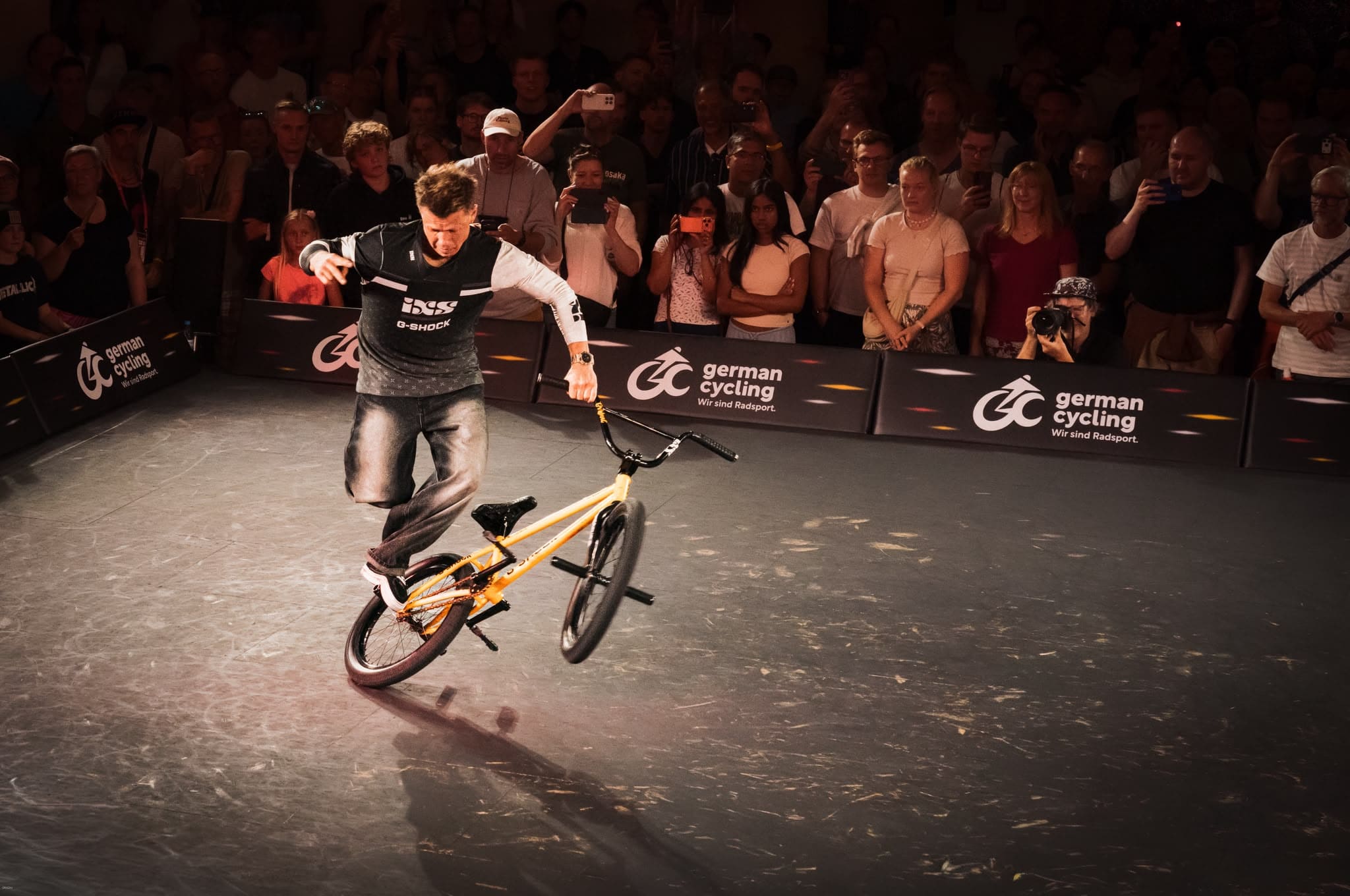 Chris Böhm macht einen BMX-Flatland-Trick auf dem Hinterrad bei den BMX Finals, gelbes Bike, ixs G-SHOCK Trikot, großes Publikum filmt und schaut zu