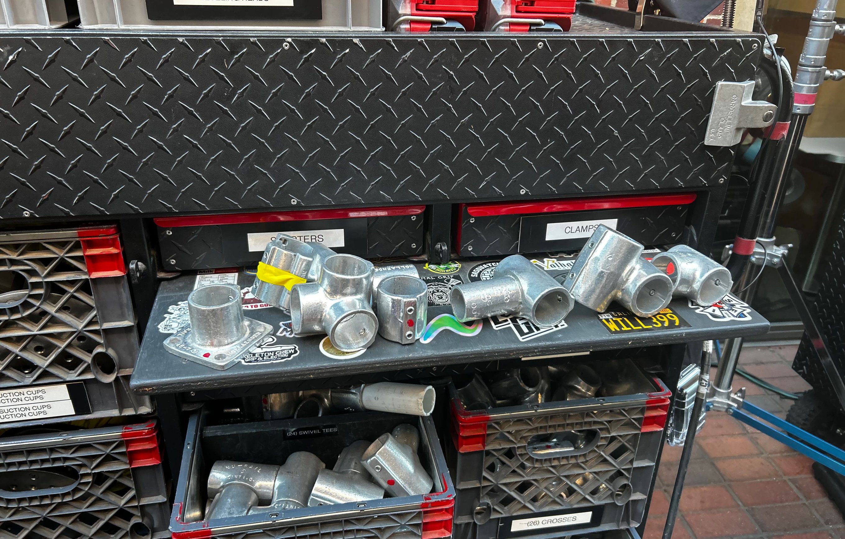 Hollaender speedrail fittings laid out on a grip-truck shelf - swivel tees, crosses, elbows and starters - above labeled crates of tees and crosses.
