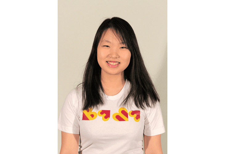 A young woman with long, dark hair and bangs stands against a light-colored background. She wears a white t-shirt with colorful, stylized heart shapes in red and yellow across the chest. Her mouth is open in a smile, and her eyes are looking directly at the camera. The image is framed by vertical teal-colored borders on either side.