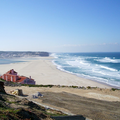 مشهد ساحلي مع الأمواج التي تتحطم على شاطئ رملي واسع، يوجد منزل بسقف أحمر على اليسار، وسماء زرقاء صافية أعلاه.