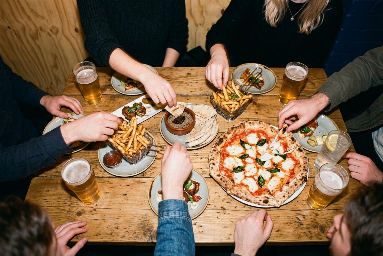Hands reaching across a wooden table crowded with a stone-baked pizza, fries, small plates and drinks, seen from above.