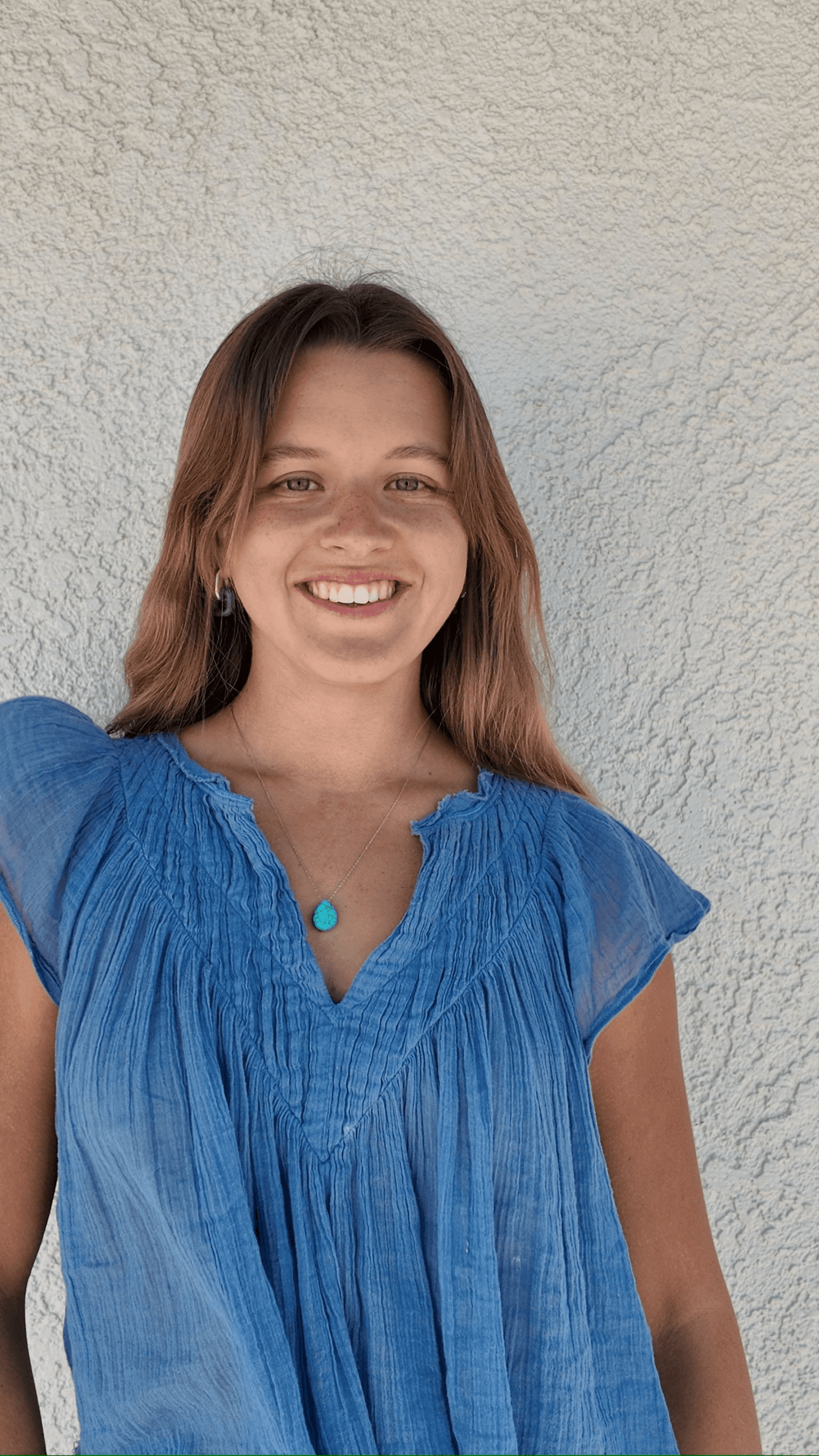 Portrait of Julia Pintar, Co-Founder of Playkit, smiling in front of a beige wall and wearing a white dress with a turquoise pendant necklace.