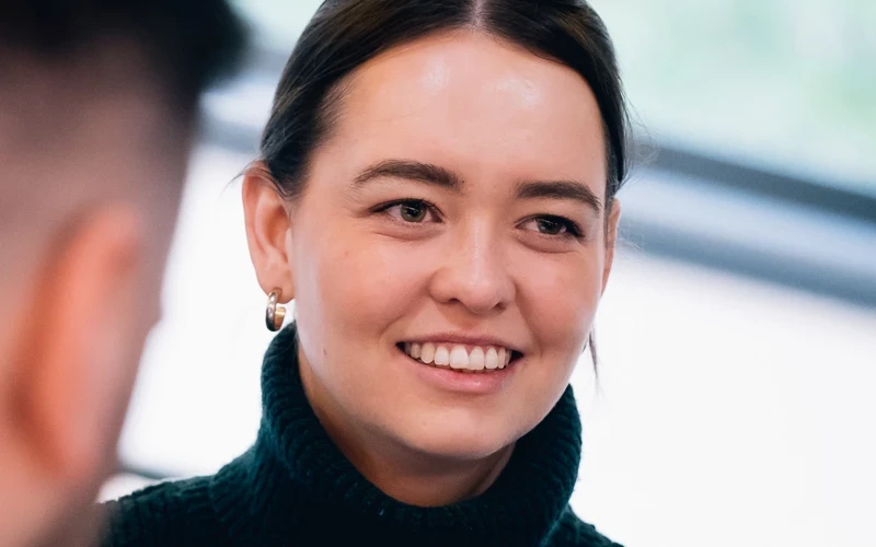 A person wearing a dark high‑neck top listens attentively during a one‑to‑one conversation indoors. The shallow depth of field focuses on the interaction, with the background softly blurred.
