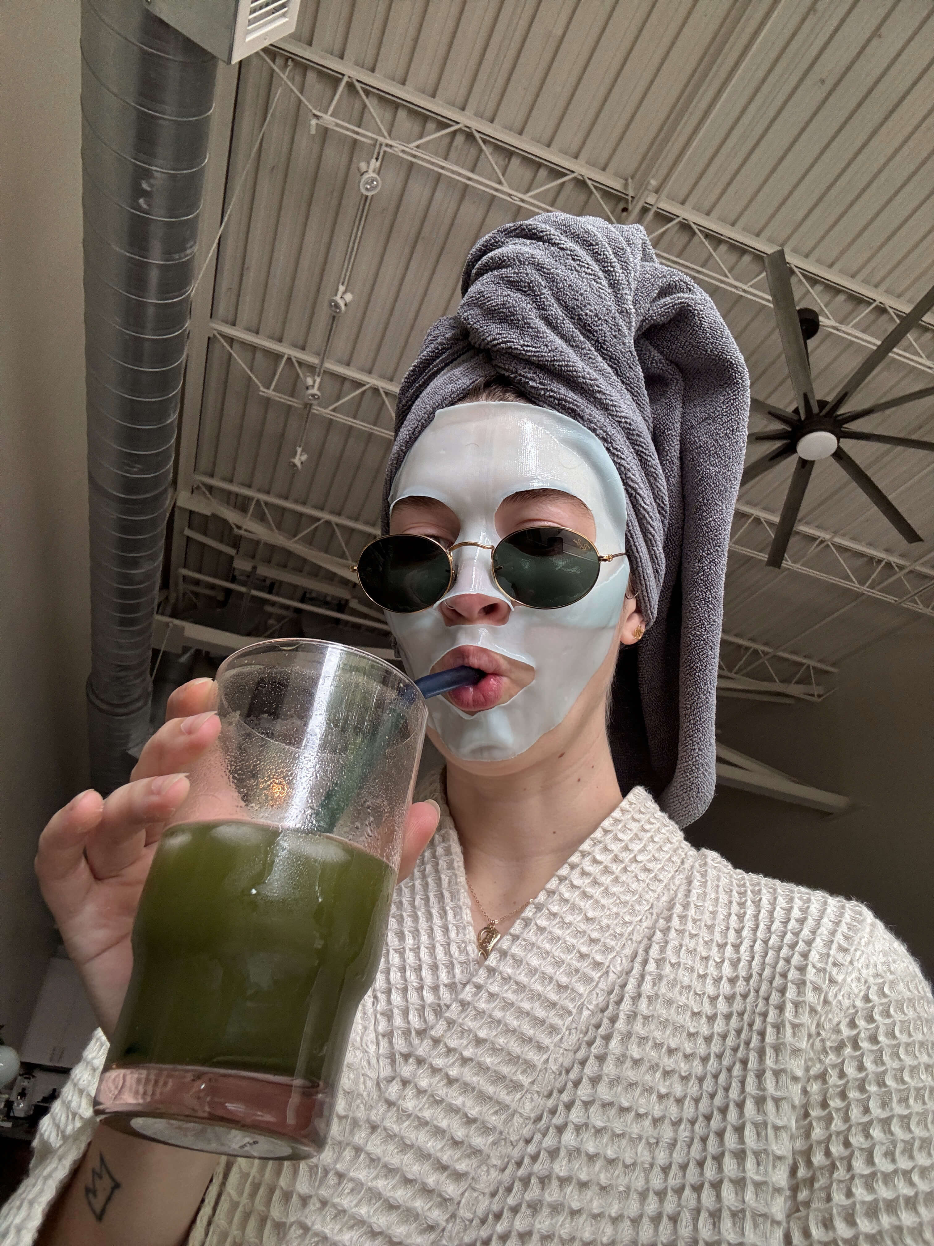 a woman wearing a robe, sunglasses, and a face mask while her hair is wrapped in a towel drinks green juice from a straw