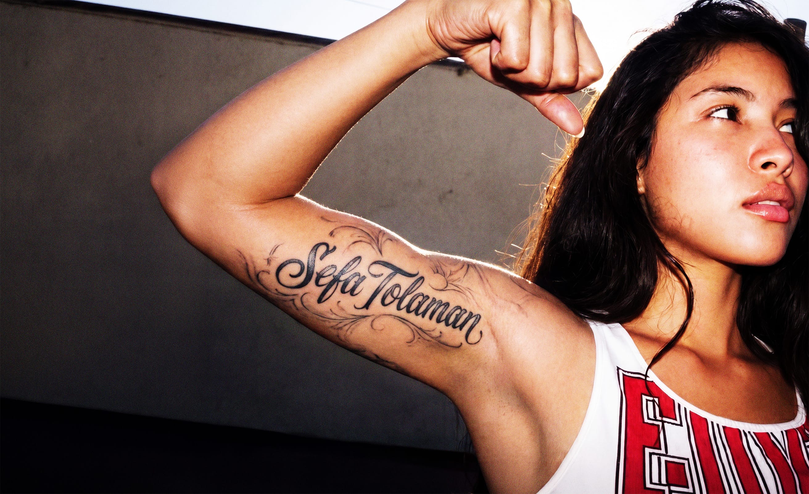 A woman with dark hair wearing a tank top and gazing up and to the right. Her left bicep has a tattoo of "Sefa Tolaman" in a stylized script.