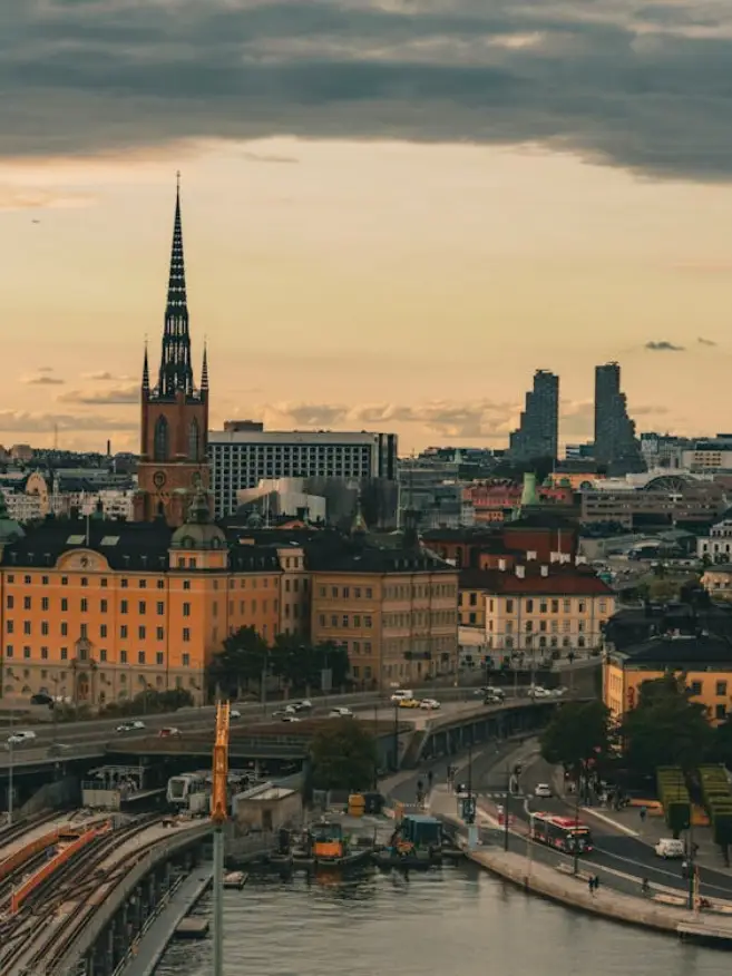 City skyline at sunset with a tall church spire, historic buildings, and modern skyscrapers. A waterway and roads lead into the bustling cityscape.