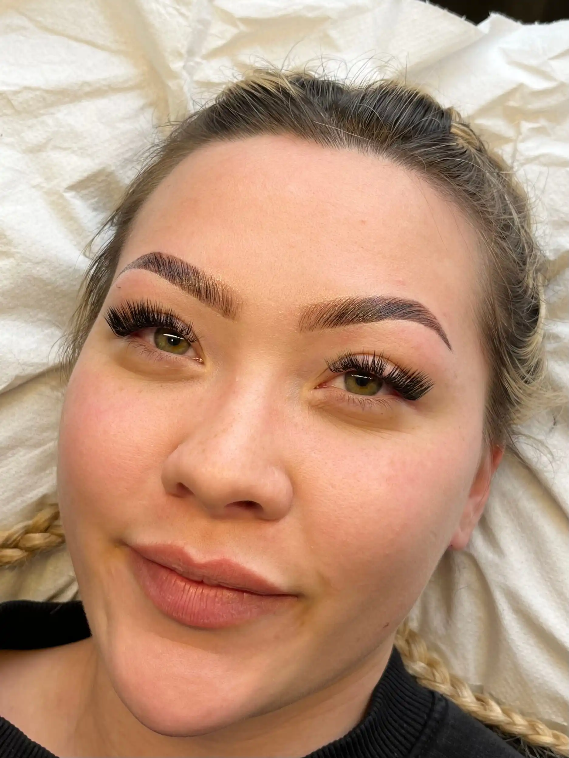 A woman with styled eyebrows and long eyelashes, lying on a white sheet. Her expression is content, conveying a sense of satisfaction and relaxation.