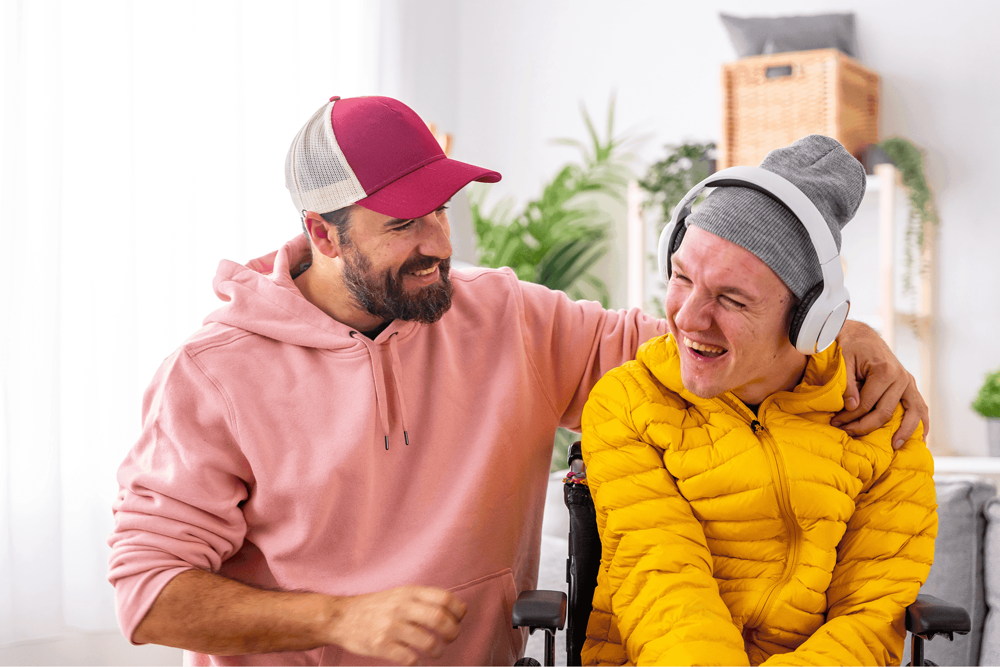 Two friends, one wearing a pink hoodie and cap and the other in a yellow jacket and gray beanie, share a joyful moment, with one listening to music on headphones in a bright living room with plants in the background.