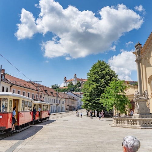 Náměstí s červenou tramvají, chodci, velkým stromem, historickými budovami a modrou oblohou s velkým mrakem.