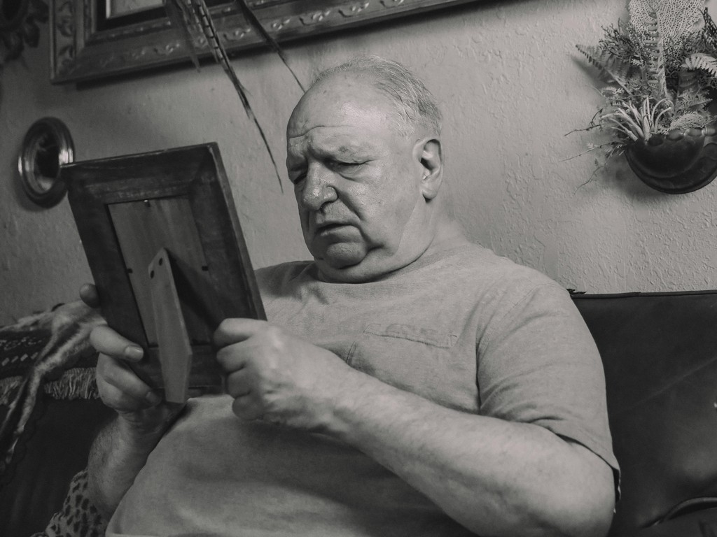 The black and white image captures an elderly man sitting on a couch, intently looking at a framed photograph he holds in his hands. His expression is contemplative and slightly somber, suggesting he is reminiscing or reflecting on memories. The setting is a cozy, lived-in room adorned with framed pictures and a decorative plant on the wall, adding to the nostalgic and intimate atmosphere.