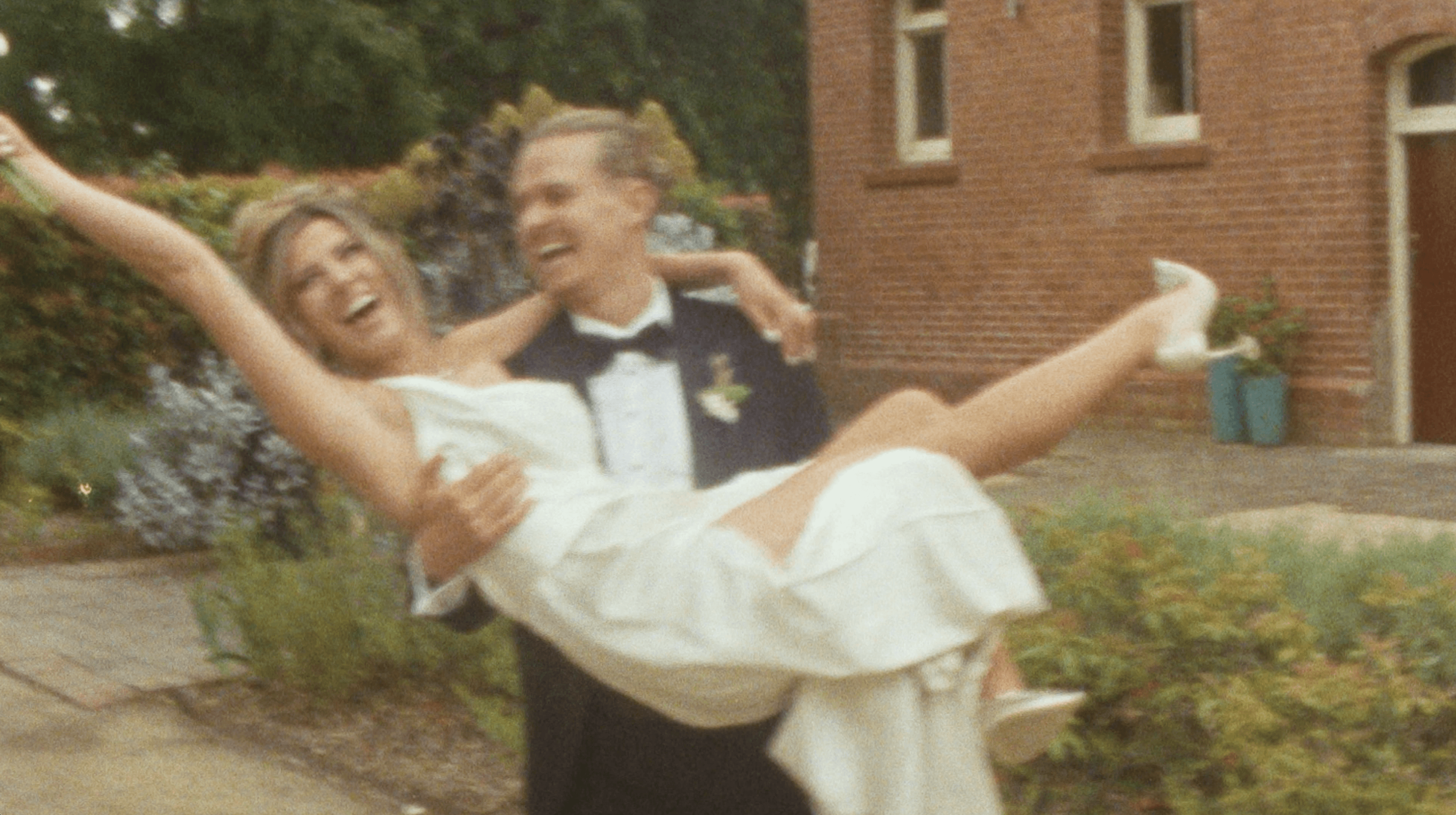A joyful couple, elegantly dressed in a white wedding gown and black tuxedo, smiles radiantly as they walk hand in hand down a lush, outdoor garden aisle, surrounded by happy guests in formal attire.