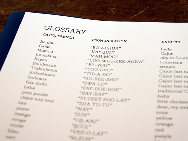 A glossary page displays French words on the left and their English translations on the right. The page is bordered by a dark blue edge. The words listed include "bonjour," "cajun," "Mamou," "Louisiana," "Bayou," "Thibodeaux," "Robicheaux," "Soileaux," "fais-dodo," "bébé," "petit poulet," "chère tout tout," "nez," "jaune," "orange," "rogo," "violet," "blue," "bonzur," "KAY-JUN," "MAH-Moo," "LOO-WEE-ZEE-ANNA," "BY-YOU," "BOO-DRO," "TIB-A-DO," "ROO-BEE-SHO," "SWA-LO," "FAY-DOE-DOE," "PI-TET FOO-LAY," "SHA TU-TU," "NAY," "JUN," "OR-ANF," "BOOF," "VEE-O-LAY," and "BLUE." The page is angled slightly and rests on a dark brown wooden surface.