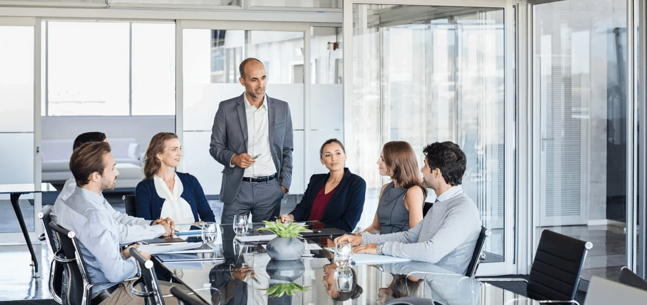 Geschäftsleute in einem modernen Konferenzraum während einer Besprechung, ein Mann steht und präsentiert vor einem Team am Tisch.