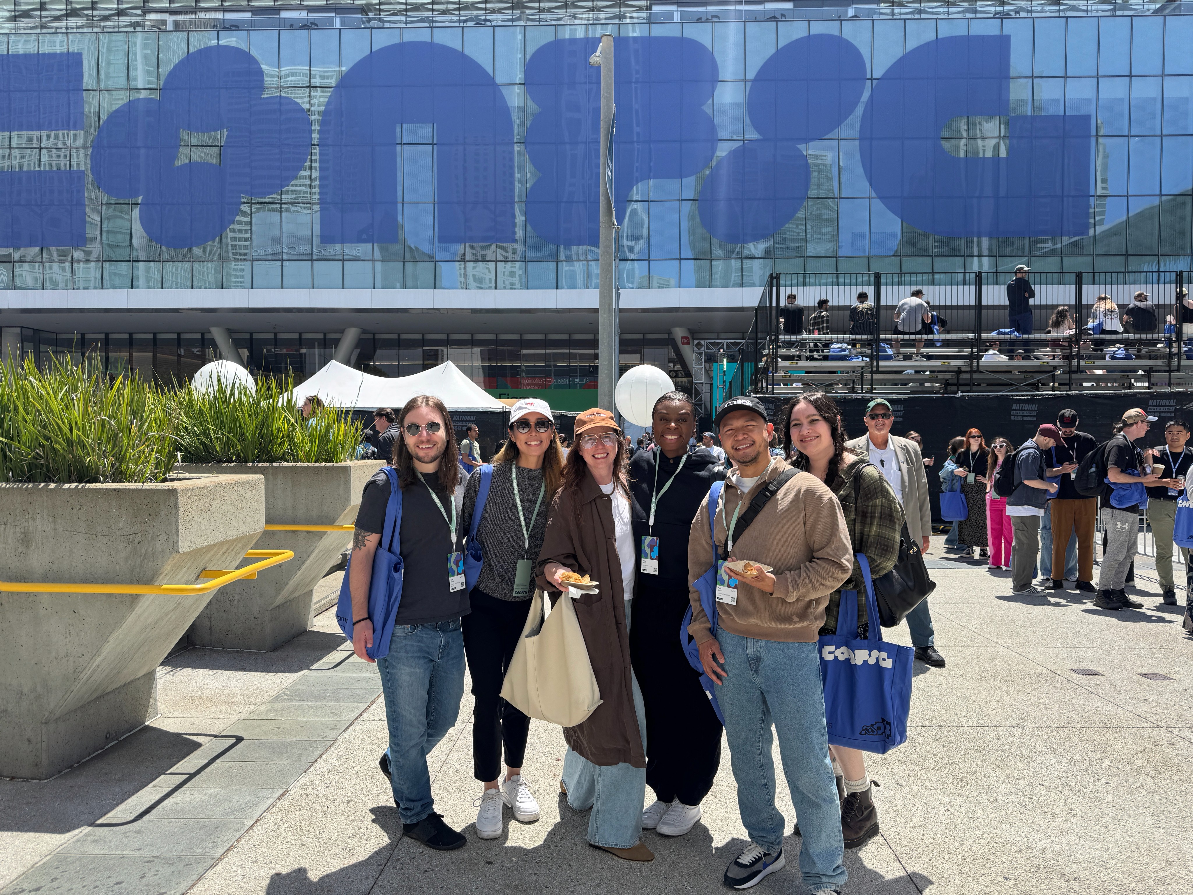 Group of people posing together outdoors in front of a large building with bold blue lettering during a daytime event.
