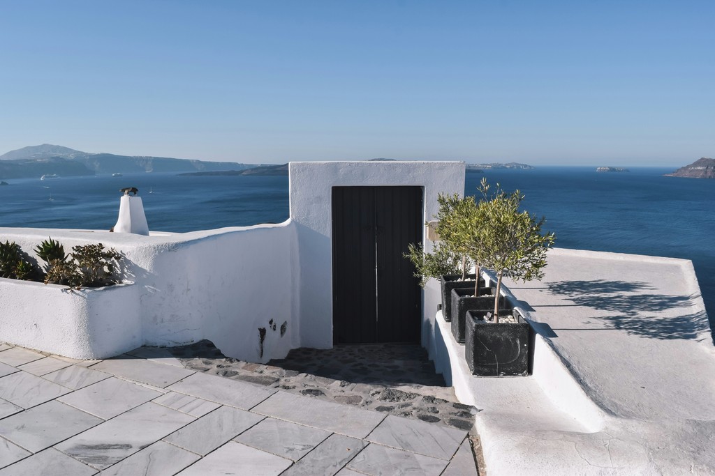 A picturesque view of a whitewashed terrace overlooking the serene blue waters and distant islands of Santorini, Greece, featuring potted olive trees and a traditional black door under a clear sky.