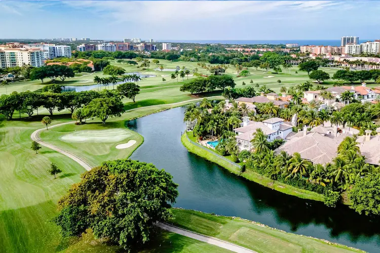 A breathtaking aerial view of a lush golf course in Boca Raton, Florida, featuring manicured greens, winding water features, and luxurious waterfront estates surrounded by palm trees. In the background, upscale residential buildings and the ocean skyline complete the picturesque scene.