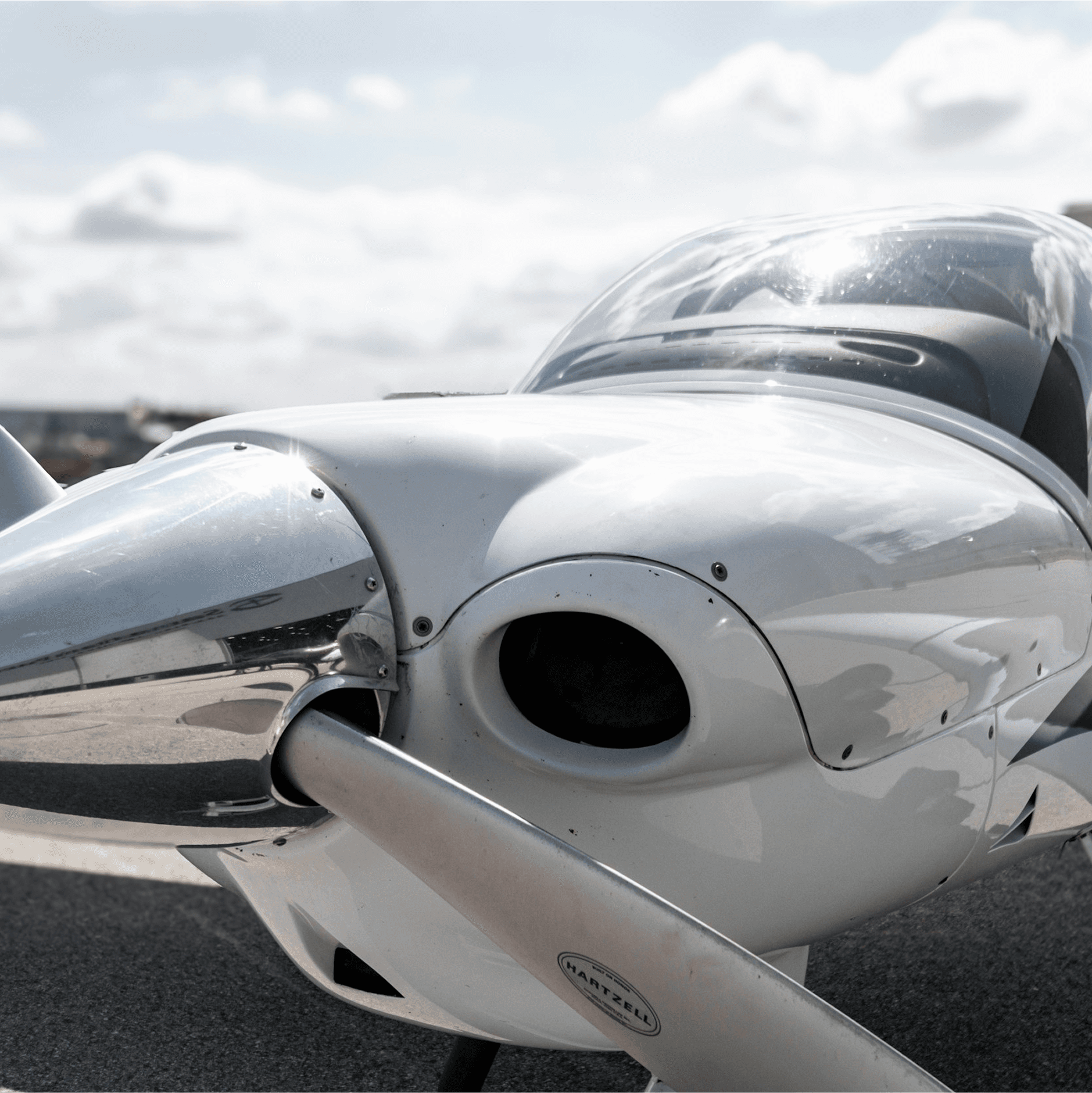 A sleek, silver propeller nose of a small, modern aircraft shines under a partly cloudy sky, with the glint of sunlight reflecting off its polished surface.