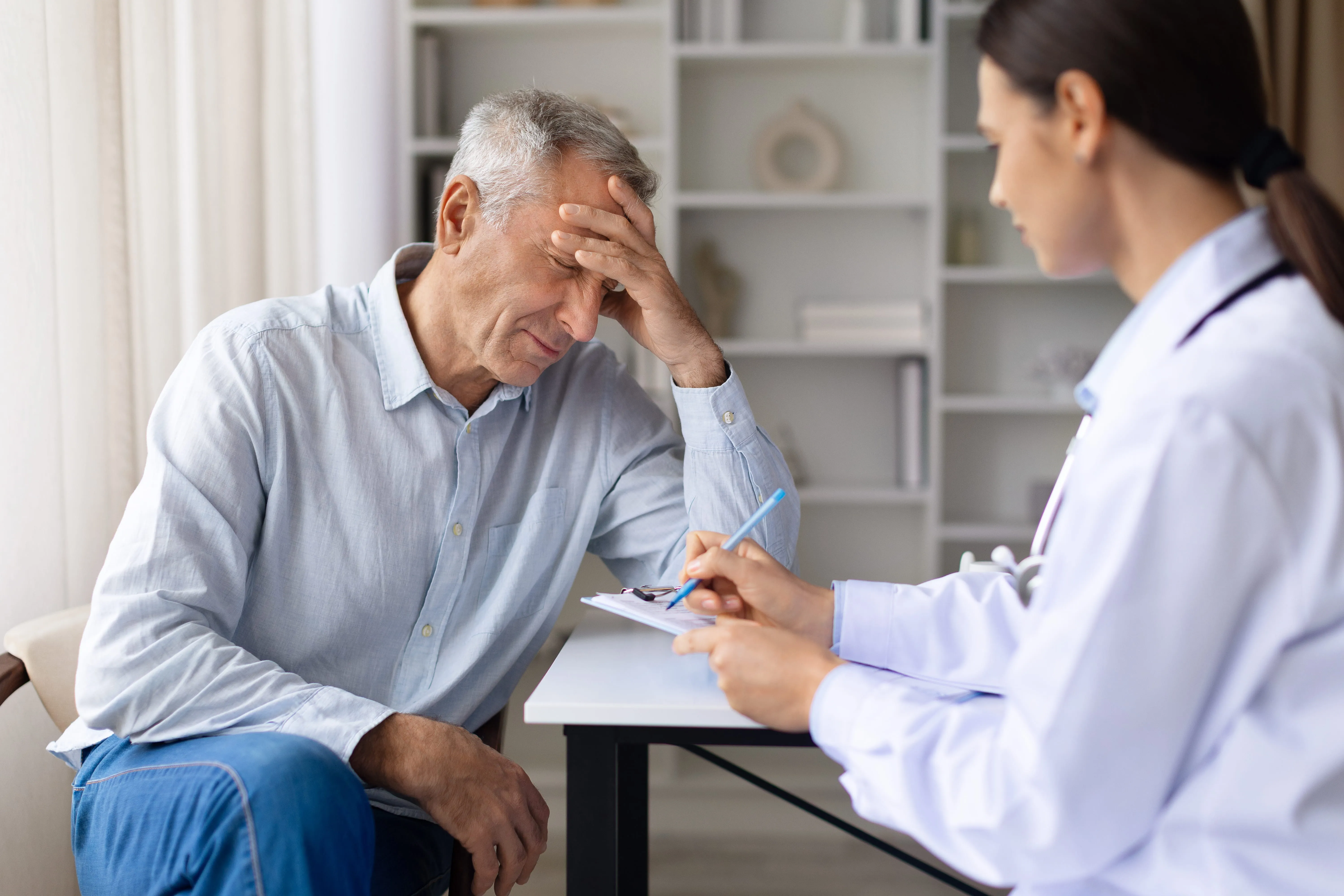 Older man holding his head in discomfort while a doctor takes notes on a clipboard during a consultation