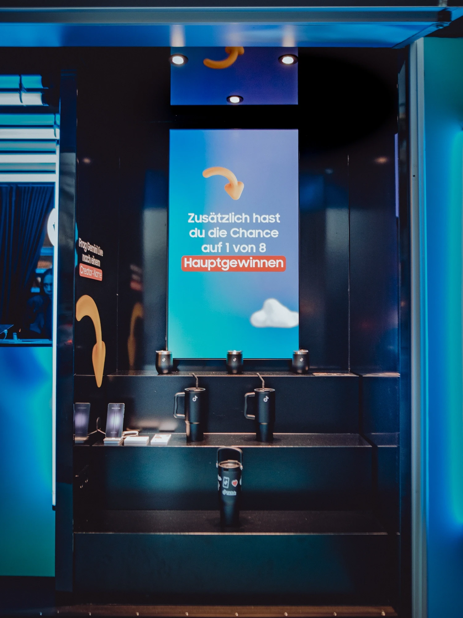 Interior of the SAMSUNG x TIKTOK module with a screen showing prize messaging and black Samsung tumbler mugs displayed on dark steps.