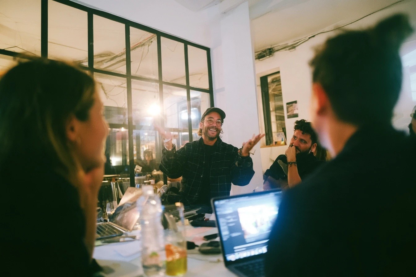 KI-Workshop bei nøho studio: Teilnehmer lachen am Tisch mit Laptops, ein Trainer erklärt mit offenen Händen