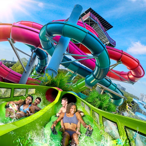 Четирима души се спускат по зелена водна пързалка с преплетени червени, сини и лилави пързалки на заден план под ясно синьо небе.