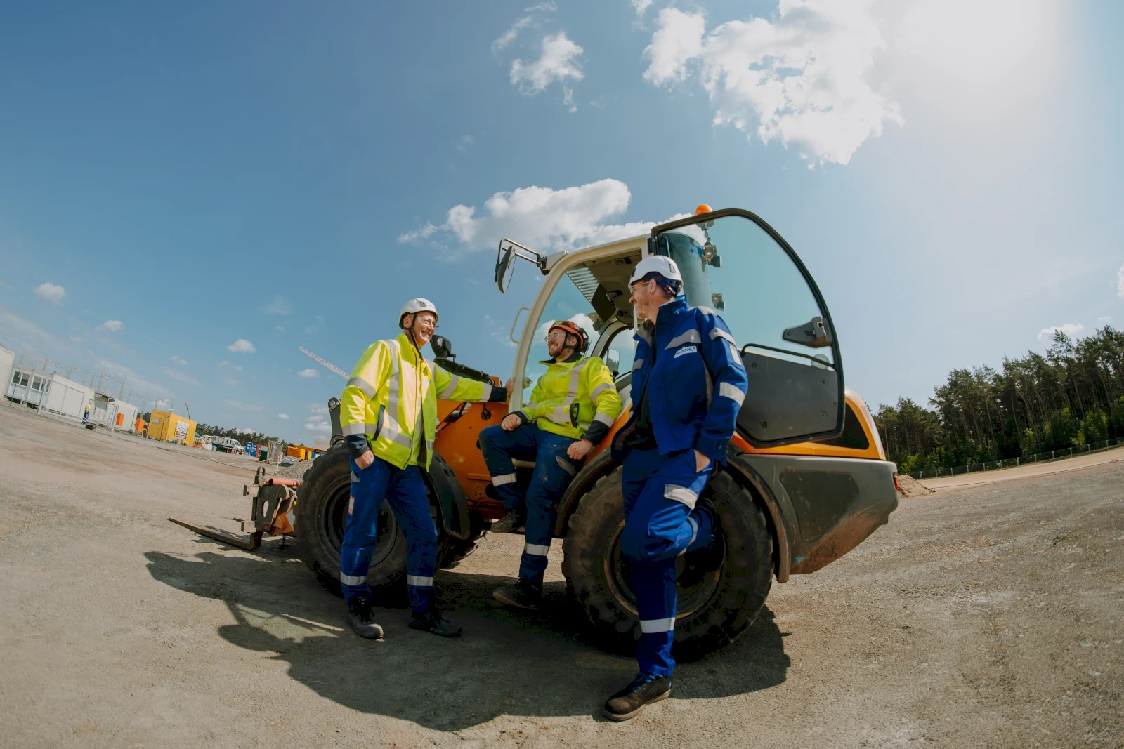 Drei Bauarbeiter stehen an einer Baumaschine und unterhalten sich bei sonnigem Wetter.