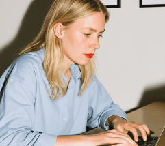A person with long blonde hair is typing on a keyboard, wearing a light blue shirt and bright red lipstick.