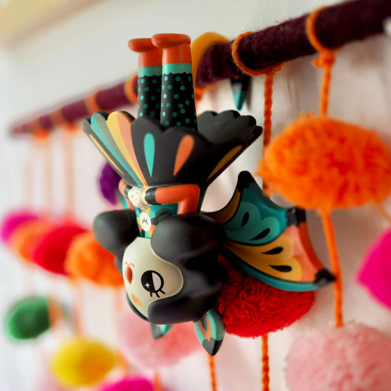 Midnight moon bat by Muxxi. Half girl, half bat, she hangs upside down for a nap. Her wings are orange yellow and blue. She's wearing a black dress with matching colour splashes, and orange boots.