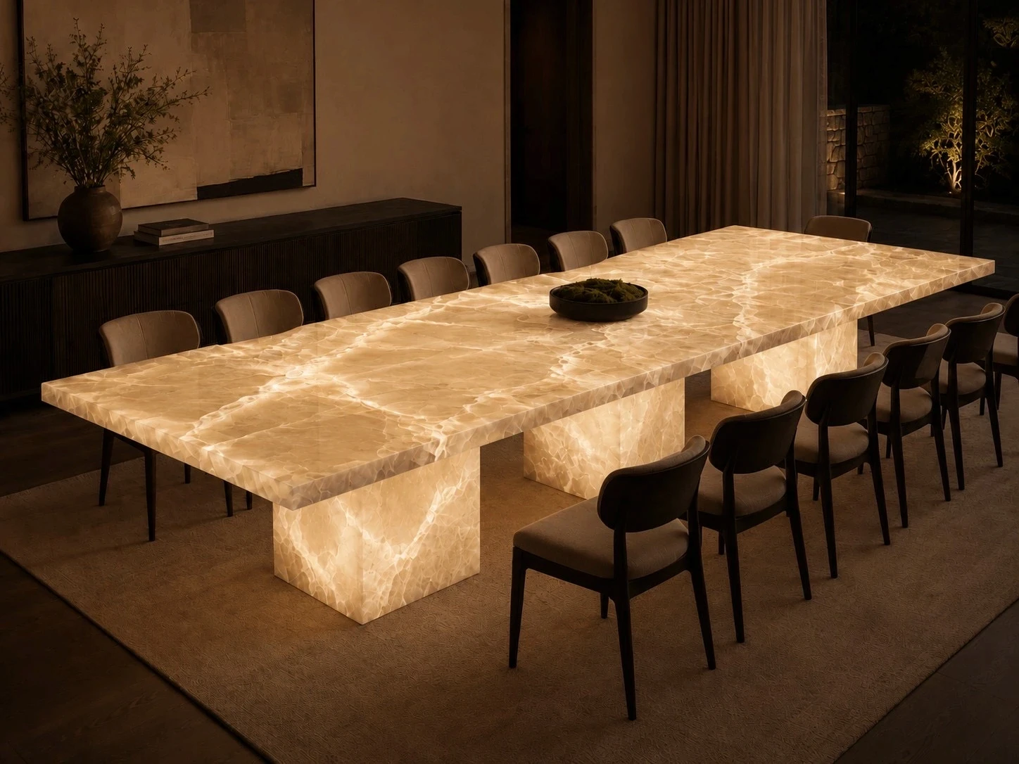 Backlit onyx dining table with a glowing translucent slab top and matching illuminated pedestal bases, surrounded by twelve taupe upholstered chairs with dark wood frames, set on a textured rug in a dimly lit dining room with a dark fluted sideboard, potted branches, abstract wall art, floor-length curtains, and a night view of a lit garden through the window.