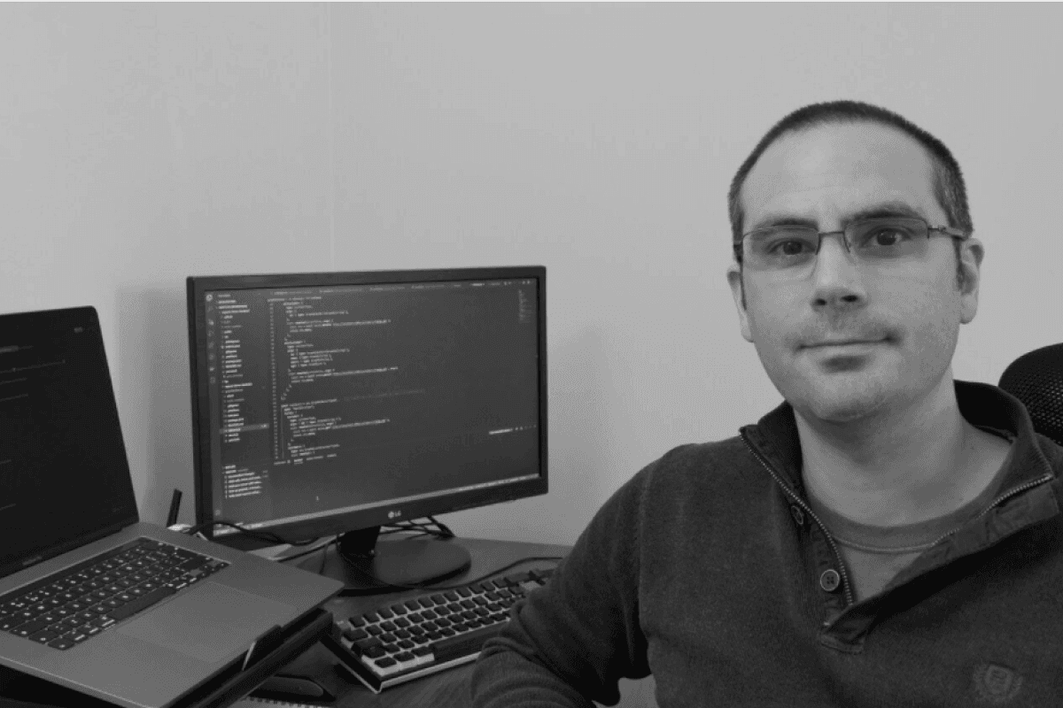 Jamie, sat at his desk smiling. There's a monitor displaying a code editor