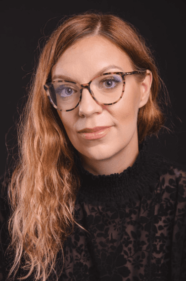 Zdjęcie przedstawia kobietę o jasnych, falowanych włosach i okularach w ciemnych oprawkach. Ma na sobie czarną bluzkę z wzorem i patrzy prosto w obiektyw. Tło jest jednolite, ciemne.