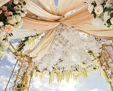 A decorative arch adorned with flowers and draped fabric, set against a bright sky.