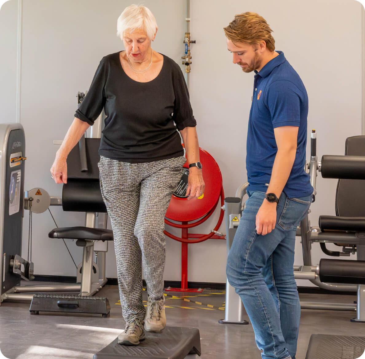 An older woman with white hair is performing a step exercise in a physical therapy clinic, guided by a young therapist wearing a blue shirt and jeans, amidst fitness equipment.