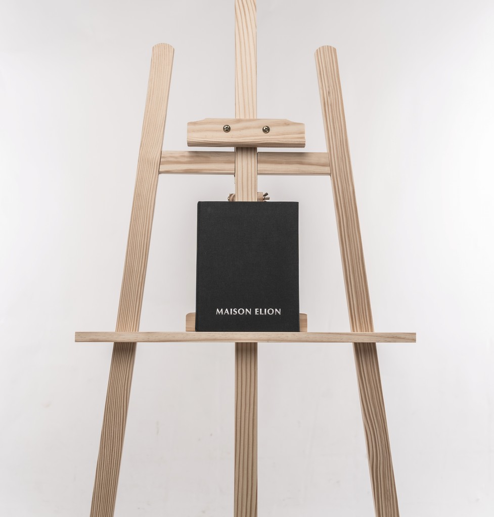 Black Fine Art album embossed “MAISON ELION”, centered on a minimalist wooden easel — purity, silence, and presence incarnate. Album Fine Art noir embossé “MAISON ELION”, centré sur un chevalet en bois minimaliste — pureté, silence et présence incarnée.