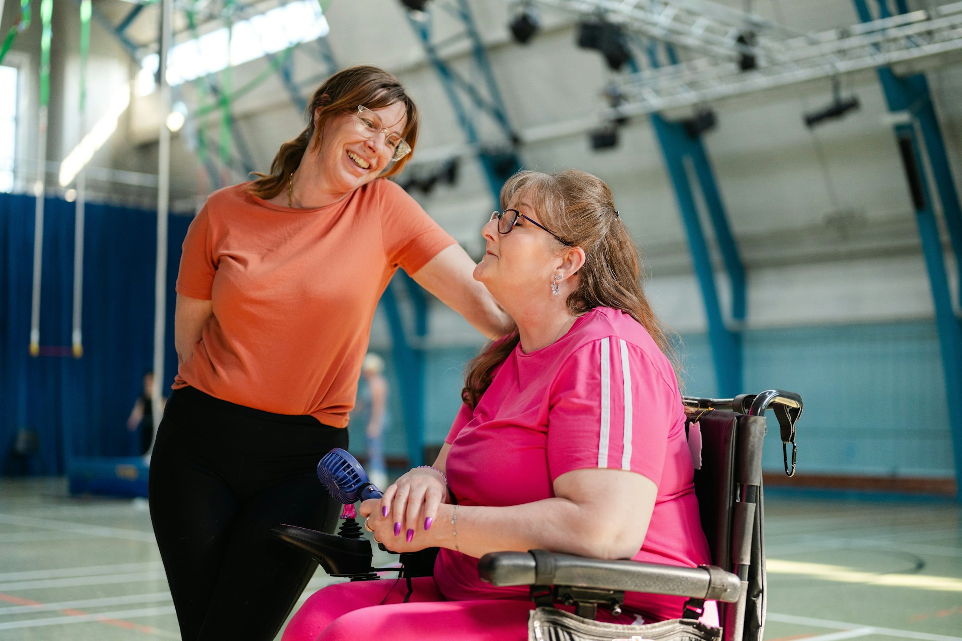 Two women smiling and talking in a sports hall, one seated in a wheelchair.