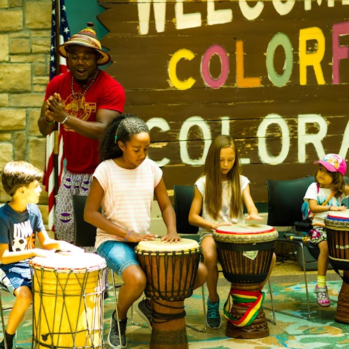 Children play drums with an instructor in front of a "Welcome to Colorful Colorado" sign.