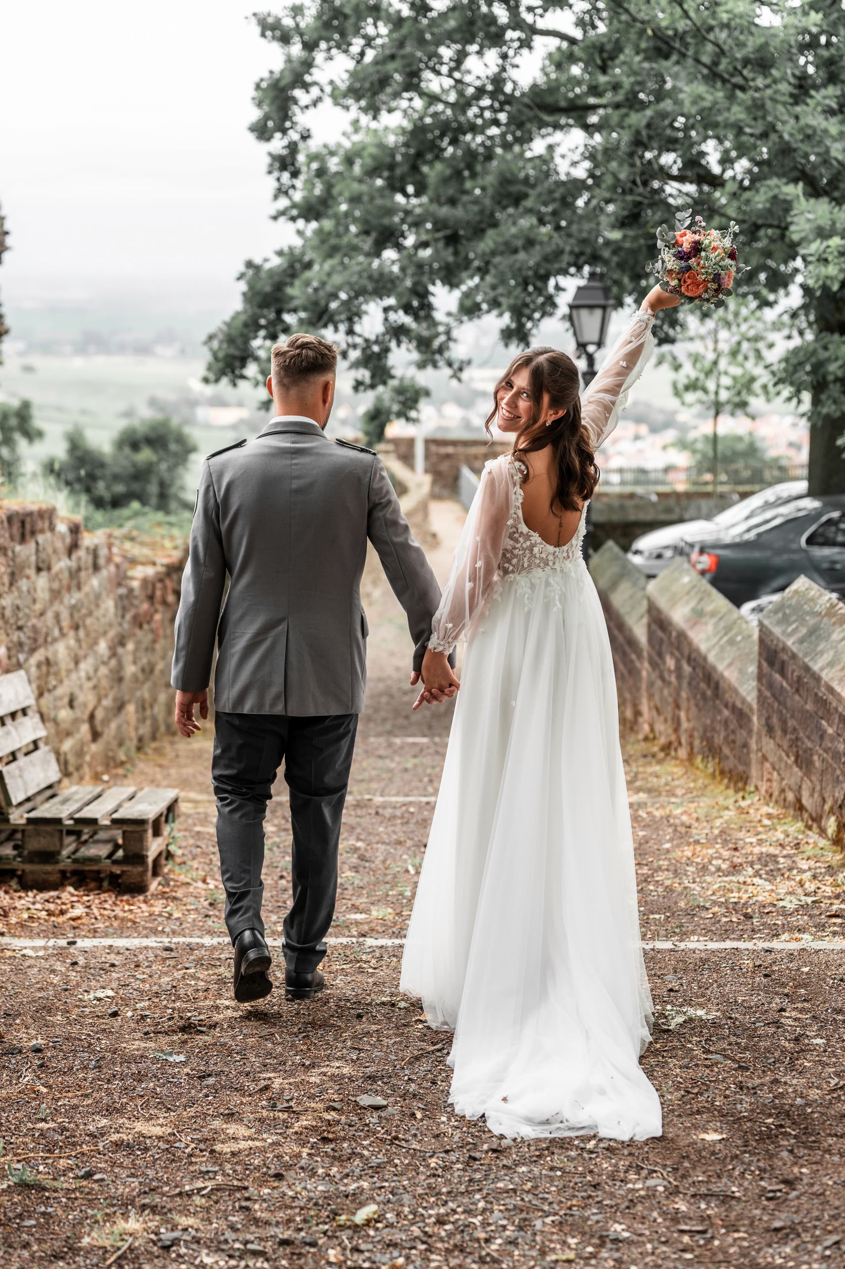 Paula und Bastian gehen Hand in Hand an der Burgmauer entlang, Paula hält den Strauß in die Höhe