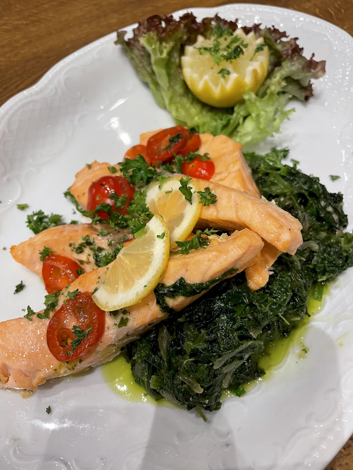 Gebratenes Lachsfilet mit Spinat, Cherrytomaten, Zitrone und Petersilie auf einem weißen Teller.